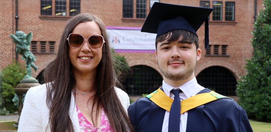 Female wearing pink dress and sunglasses standing with male wearing graduation cap and gown over shirt and tie.